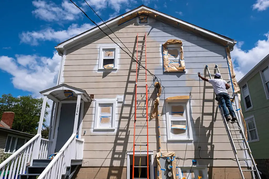 Professional painter applying fresh exterior coating on a two-story home, showing how weather affects exterior paint longevity in seasonal climates