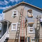 Professional painter applying fresh exterior coating on a two-story home, showing how weather affects exterior paint longevity in seasonal climates