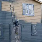 Painter on a ladder spraying exterior siding on a two-story home in Warwick, NY, with windows masked and ready for an exterior painting project.