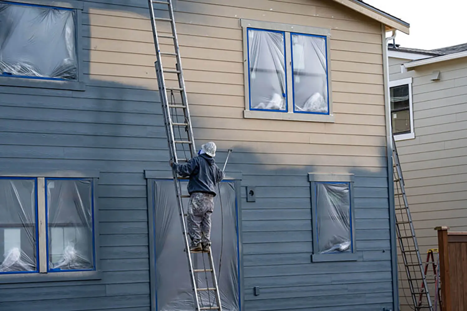 Painter on a ladder spraying exterior siding on a two-story home in Warwick, NY, with windows masked and ready for an exterior painting project.