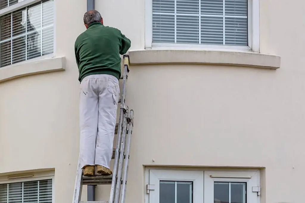 Painter on a ladder working on the exterior of a home, demonstrating how spring weather conditions affect exterior painting projects.