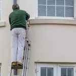 Painter on a ladder working on the exterior of a home, demonstrating how spring weather conditions affect exterior painting projects.