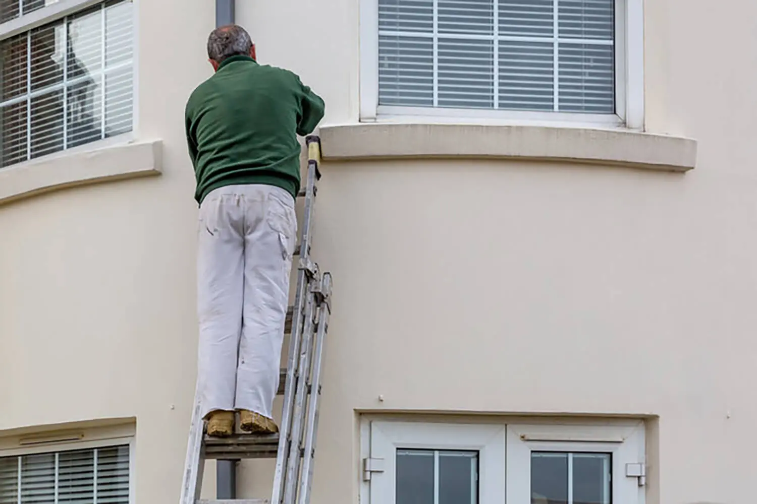 Painter on a ladder working on the exterior of a home, demonstrating how spring weather conditions affect exterior painting projects.