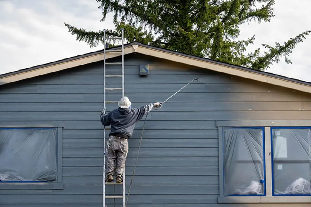 Painter on a ladder spraying exterior siding on a residential home, showing the active work involved in a summer vs. spring exterior painting project.