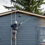 Painter on a ladder spraying exterior siding on a residential home, showing the active work involved in a summer vs. spring exterior painting project.