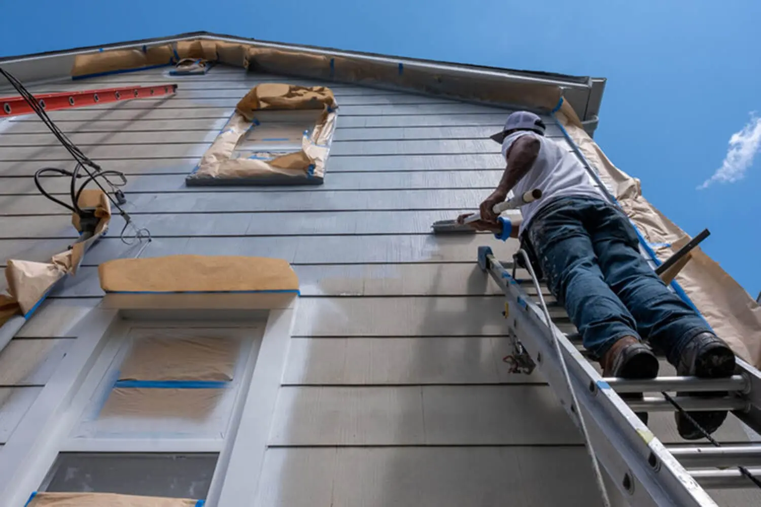 Painter on a ladder rolling exterior siding on a residential home on a sunny day, showing the pros and cons of exterior painting in summer in Warwick, NY.