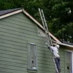 Professional painter on a ladder spraying exterior siding, illustrating how long professional exterior painting typically lasts.