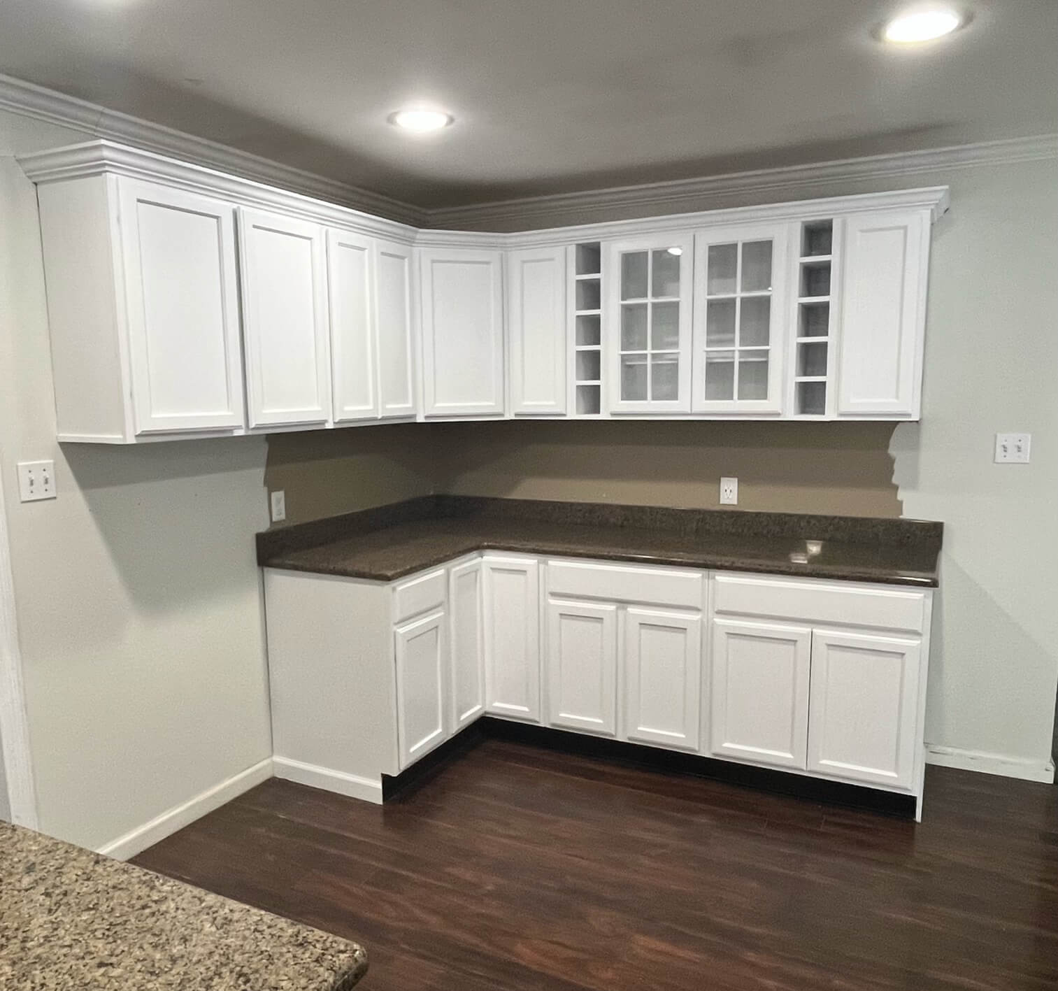 A corner view of a newly remodeled kitchen featuring white shaker cabinets, dark brown granite countertops, dark hardwood flooring, and light green walls.