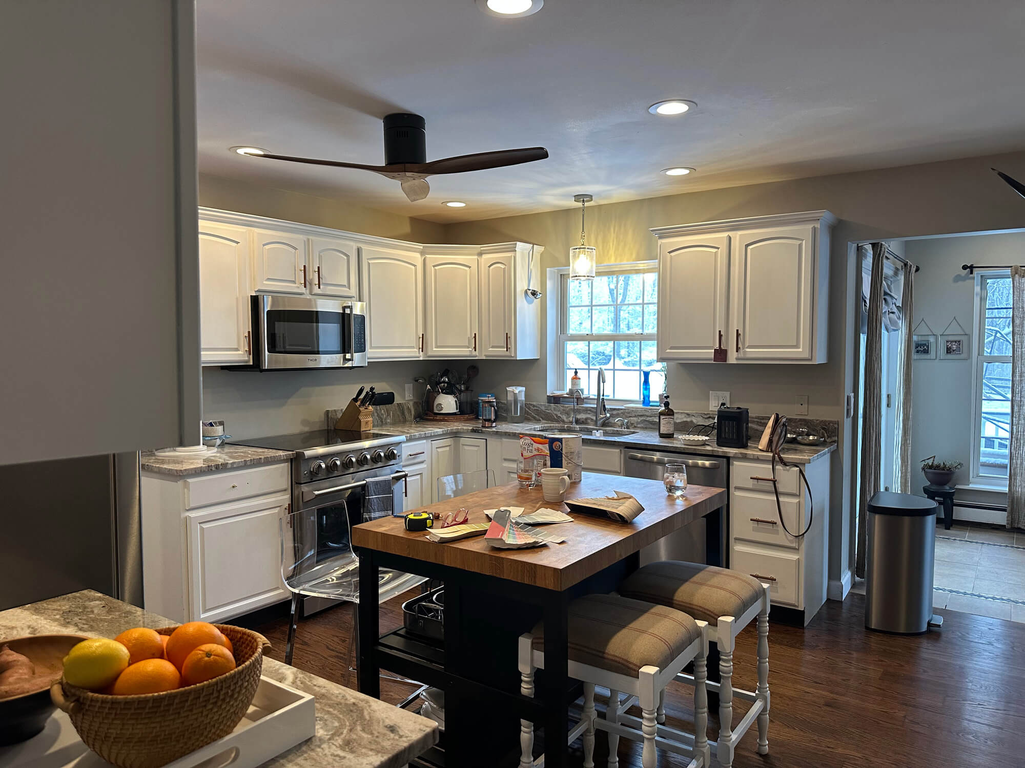 A bright, modern kitchen featuring white shaker cabinets, a stainless steel microwave and oven, a dark wood butcher block island with seating, and dark hardwood floors.