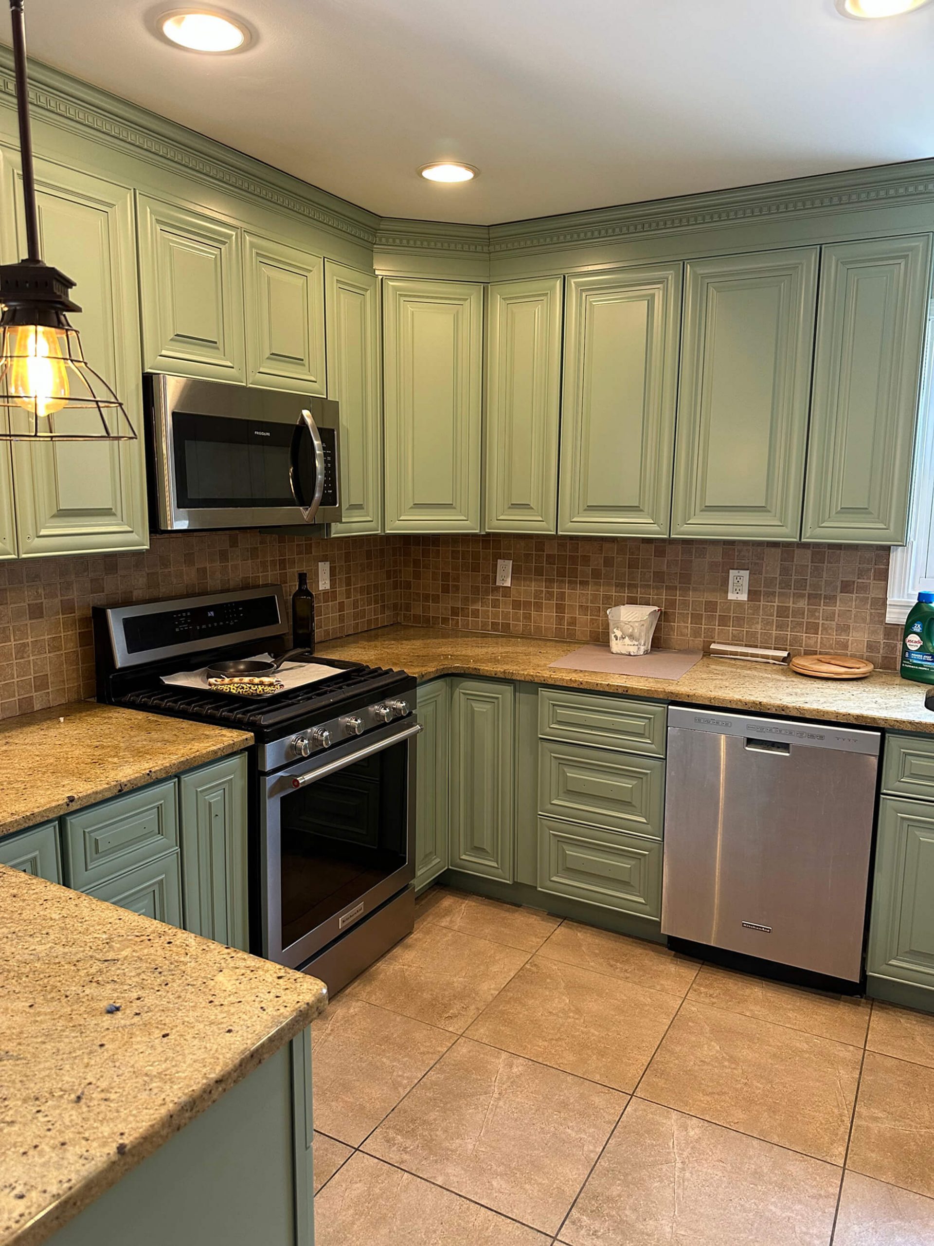 A kitchen featuring sage green cabinets, speckled beige granite countertops, stainless steel appliances including an oven and microwave, a brown and beige tiled backsplash, and light brown floor tiles.