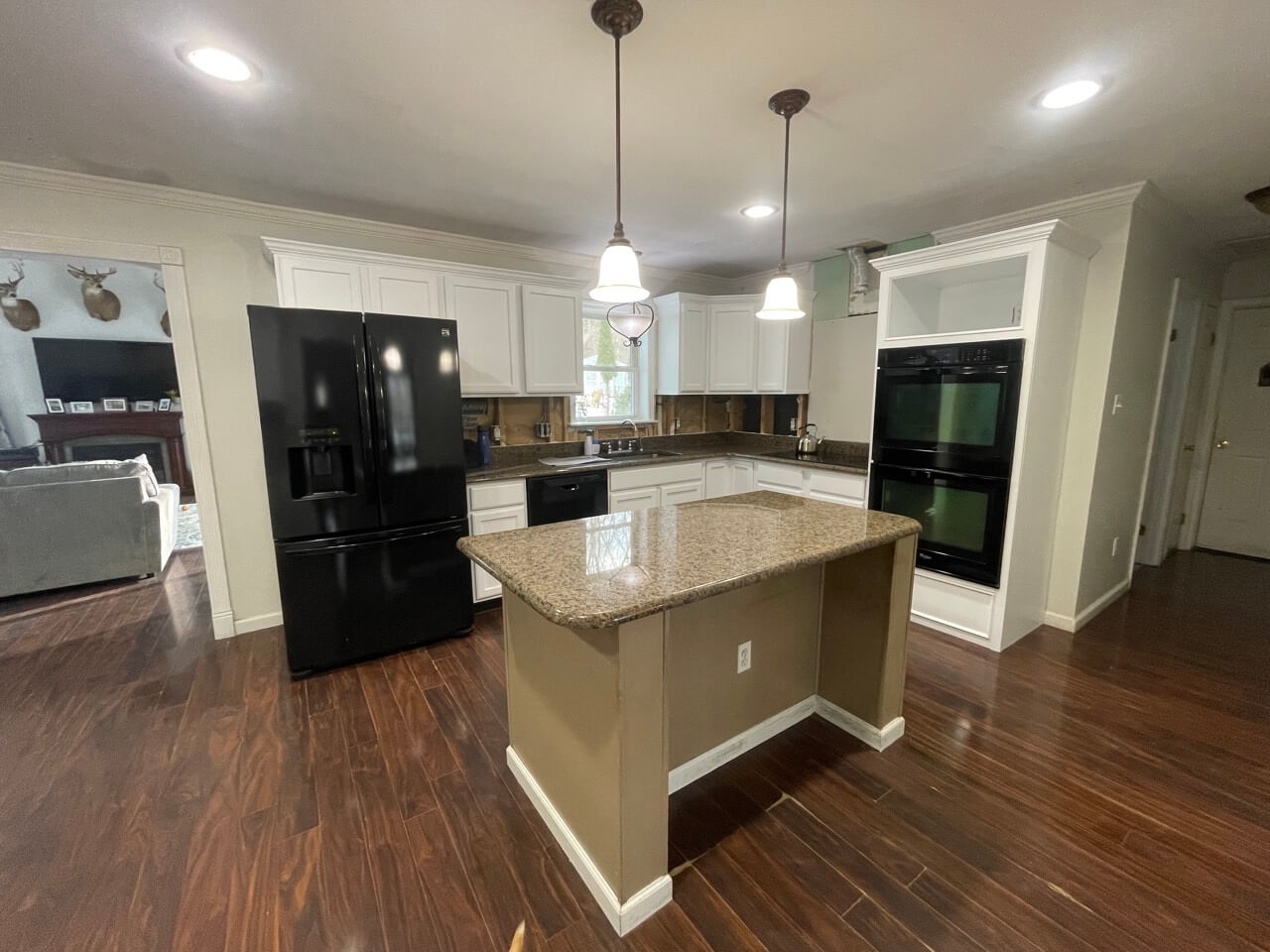 A spacious kitchen with white cabinets, dark wood floors, a black refrigerator and double oven, a beige island with granite countertop, and pendant lights hanging above it, opening to a living area in the background.