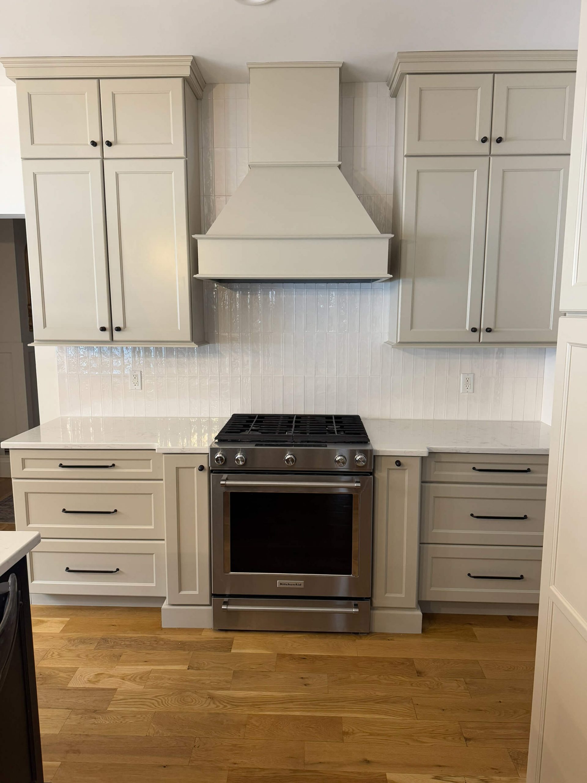 A detail shot of a modern kitchen focusing on a stainless steel gas range set between light grey cabinets, a custom hood, white quartz countertops, and a vertical subway tile backsplash.