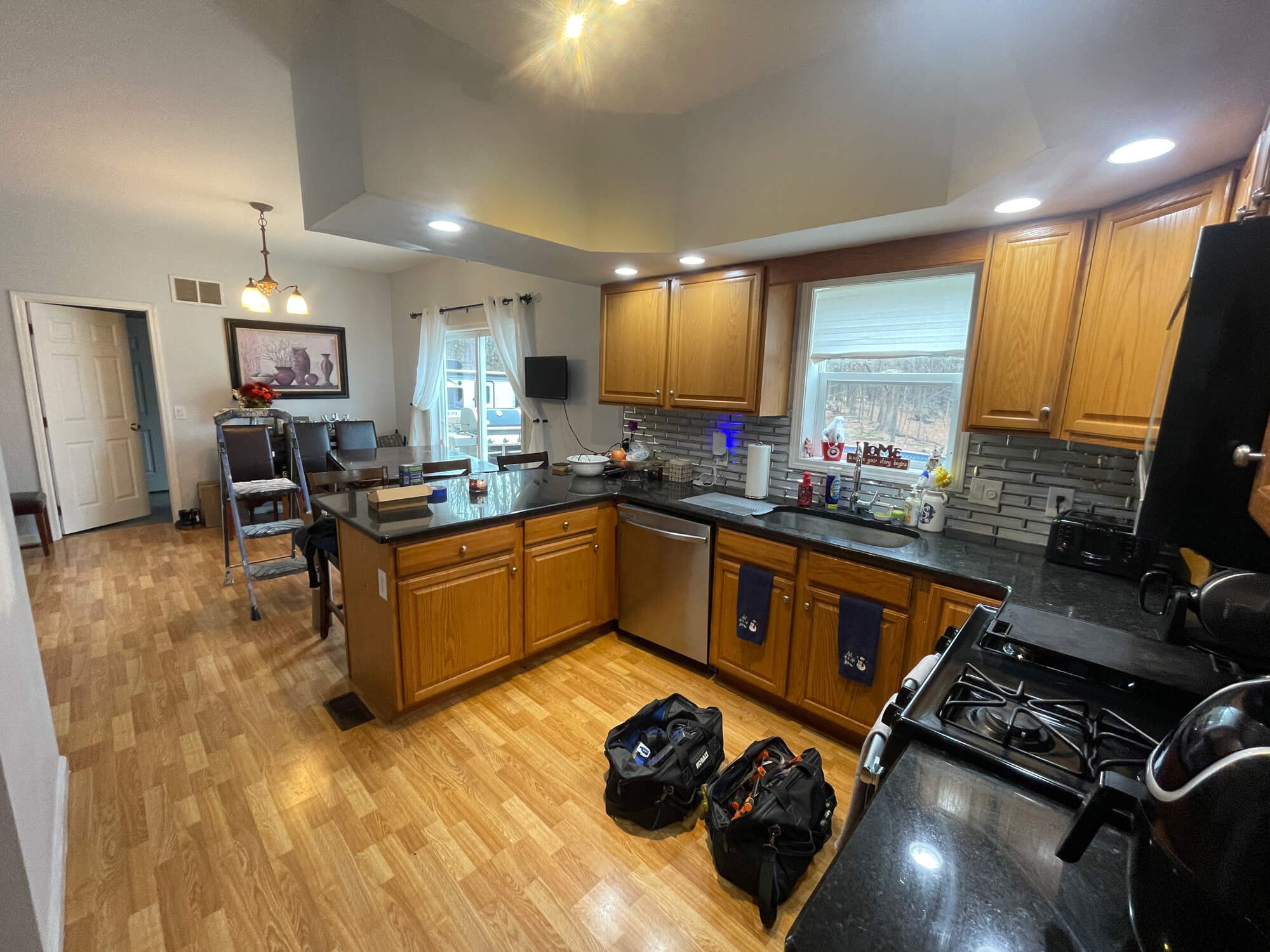 An interior view of a kitchen with honey oak cabinets, black countertops, and wood laminate flooring, showing two tool bags on the floor and an adjacent dining area.