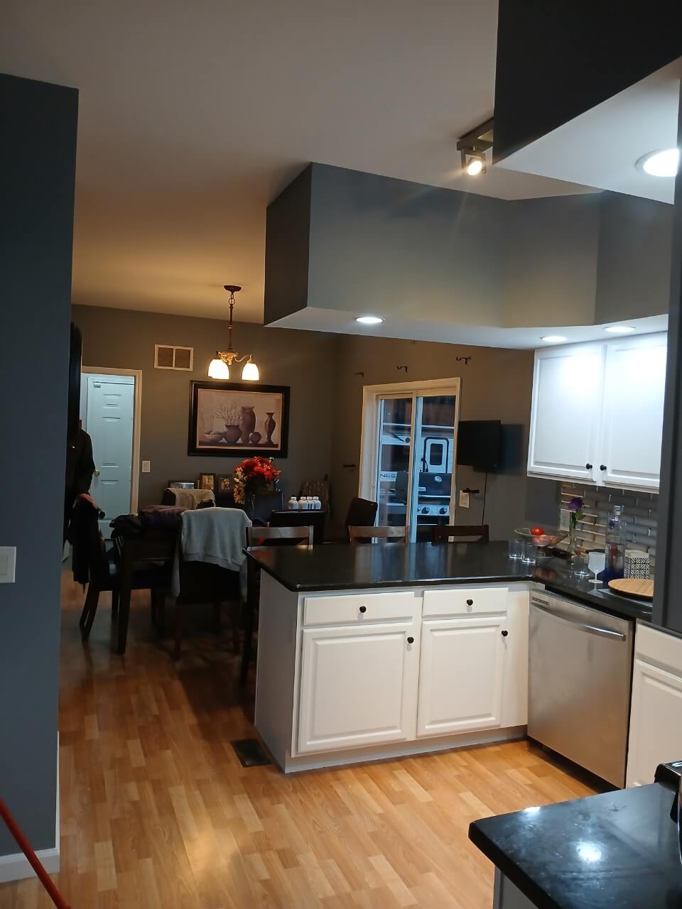 A kitchen and dining area featuring light wood floors, dark grey walls, white cabinets and island with black countertops, and a dining table visible through the entryway.