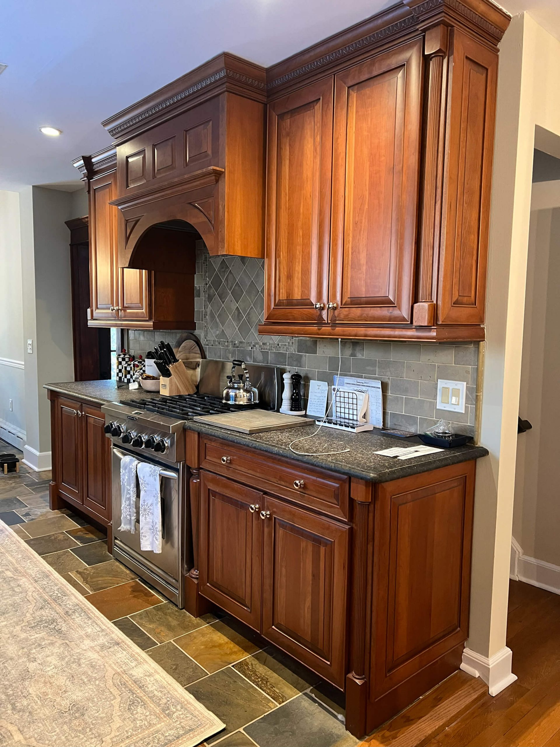 A luxurious kitchen corner featuring dark cherry wood cabinetry, a stainless steel range, dark stone countertops, and a grey diamond-patterned tile backsplash above a slate tile floor.
