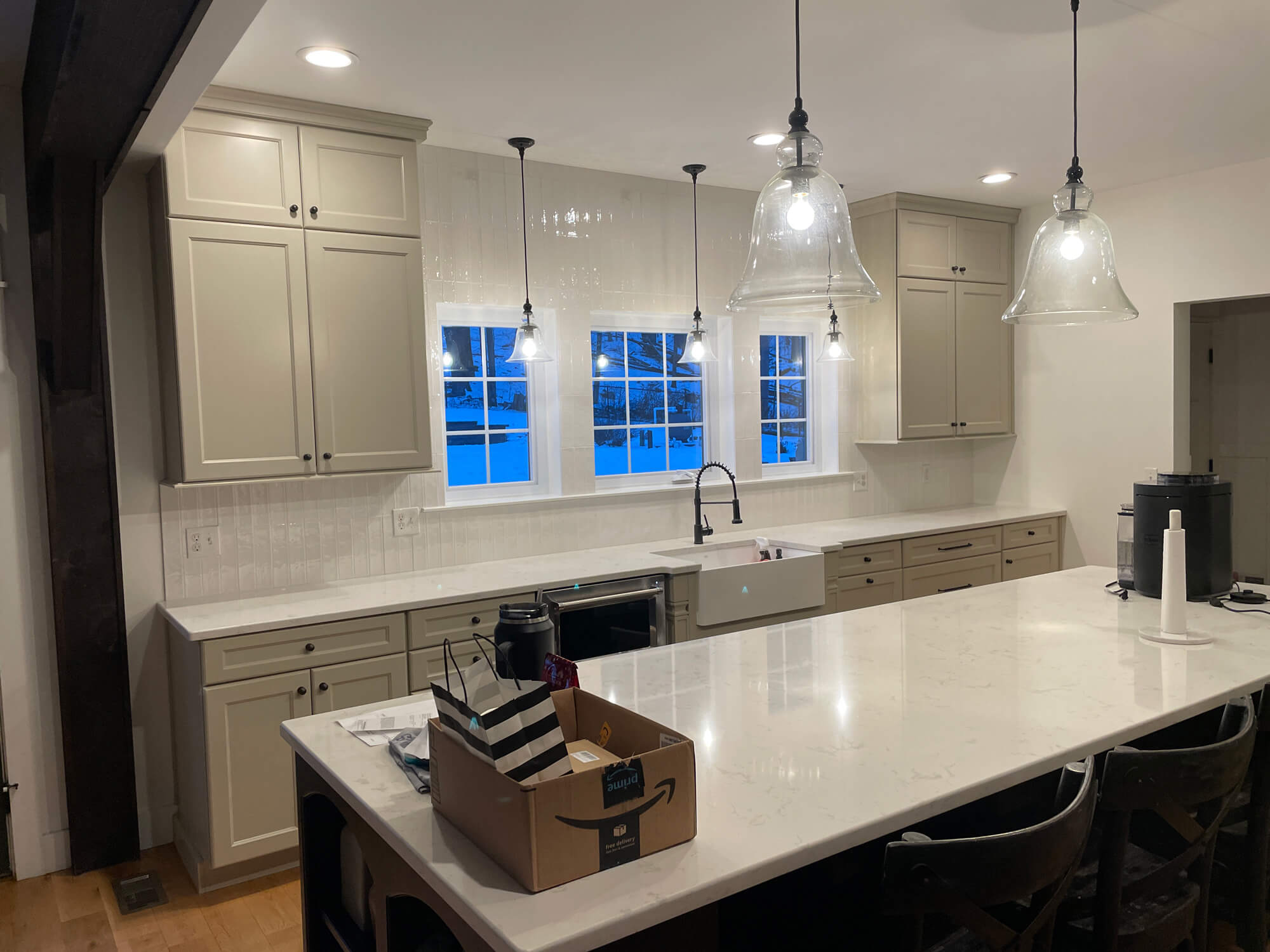 A newly renovated kitchen featuring taupe shaker cabinets, a long white island with seating, a farmhouse sink beneath three windows, and large glass pendant lights.