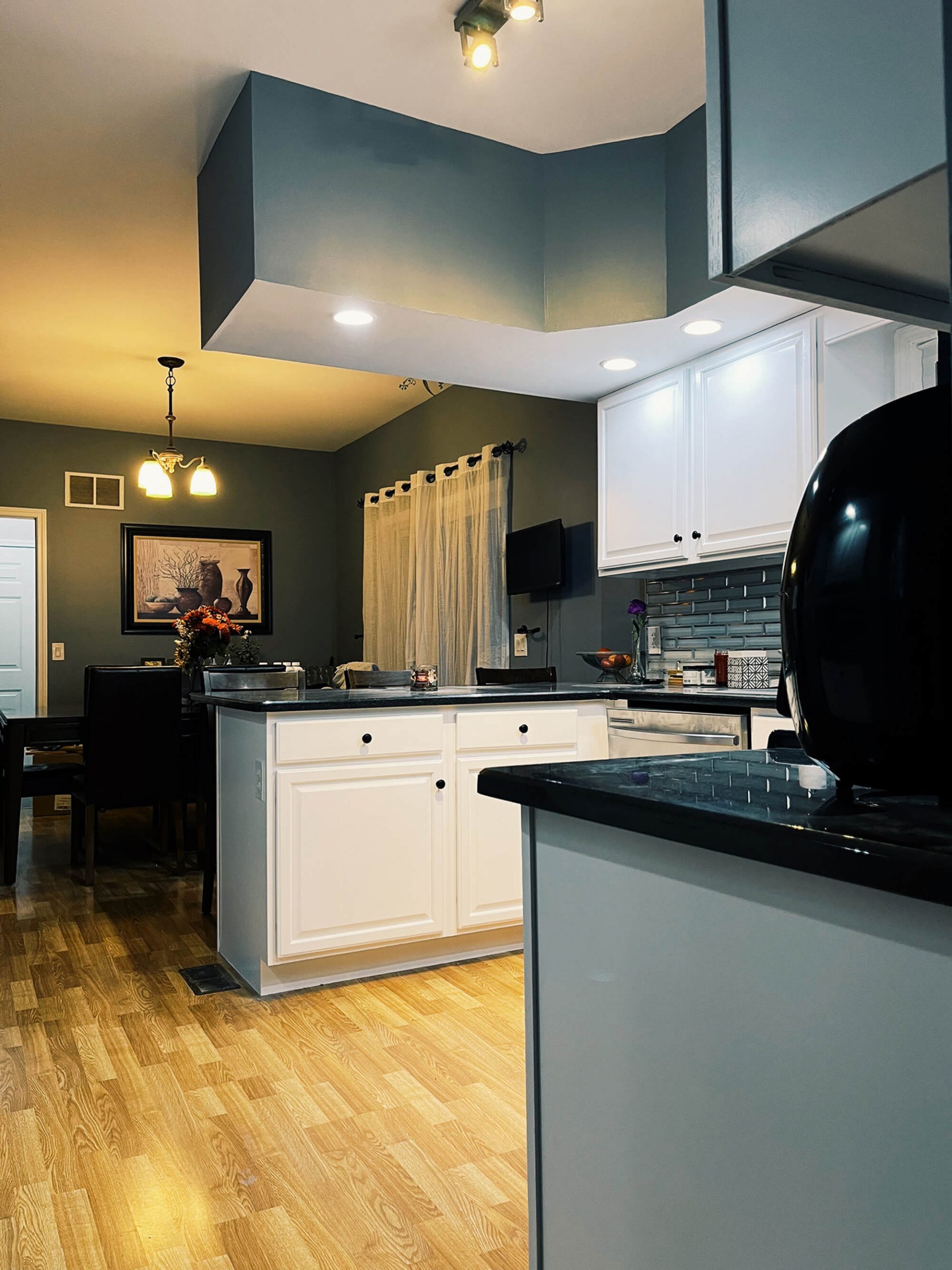 A kitchen and dining room with laminate wood flooring, dark teal walls, white cabinets and island with black countertops, and a dining table with a framed still life painting.