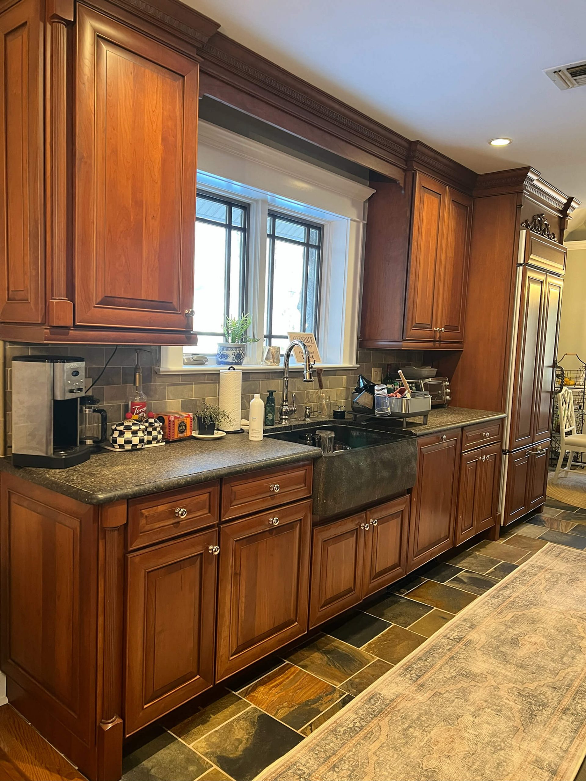 A traditional kitchen with rich cherry wood cabinets, dark countertops, a dark farmhouse sink beneath a multi-paned window, and a floor covered in dark, multi-colored slate tiles with an area rug.