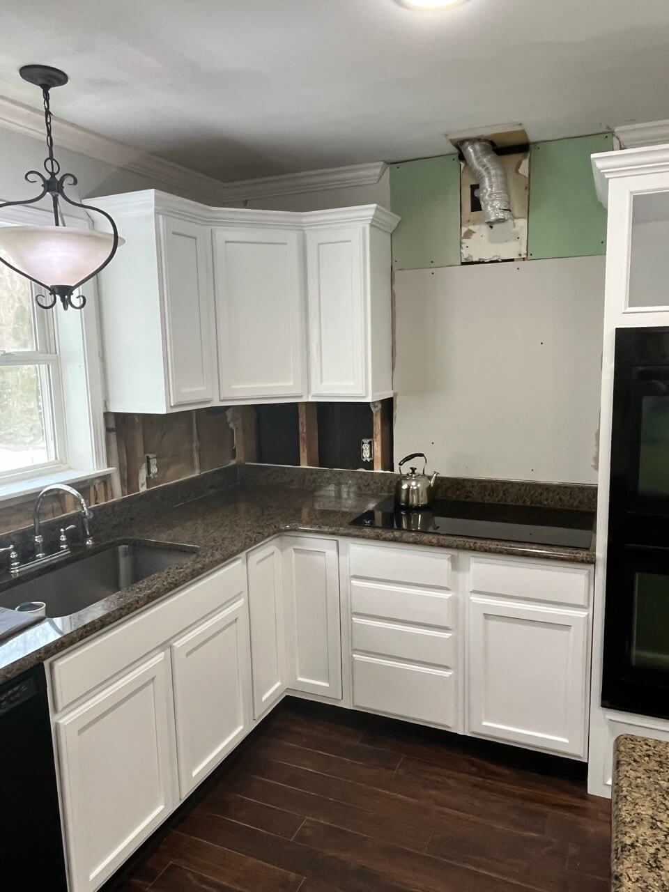 A kitchen renovation in progress featuring white cabinets, dark granite countertops, dark wood flooring, and exposed drywall where the backsplash is missing, revealing a vent pipe above.