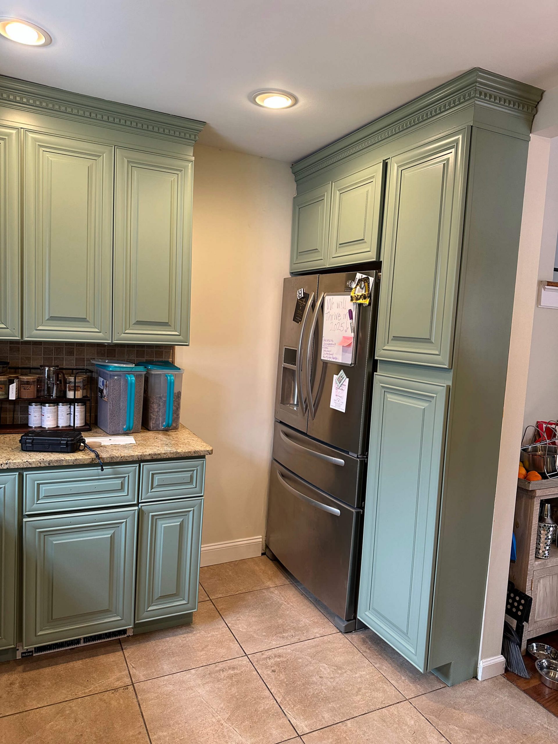 A kitchen corner featuring sage green cabinets surrounding a stainless steel French door refrigerator, speckled beige granite countertops, and large square floor tiles.
