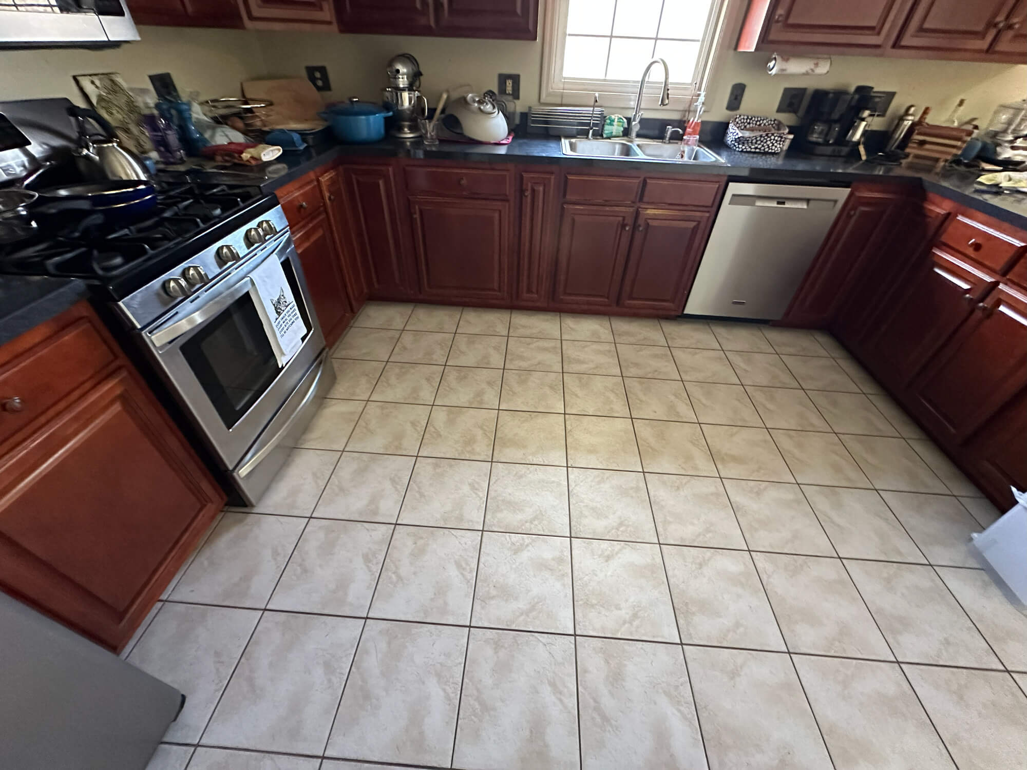 A U-shaped kitchen featuring warm cherry wood cabinets, black countertops, a stainless steel stove and dishwasher, and light beige square tile flooring.