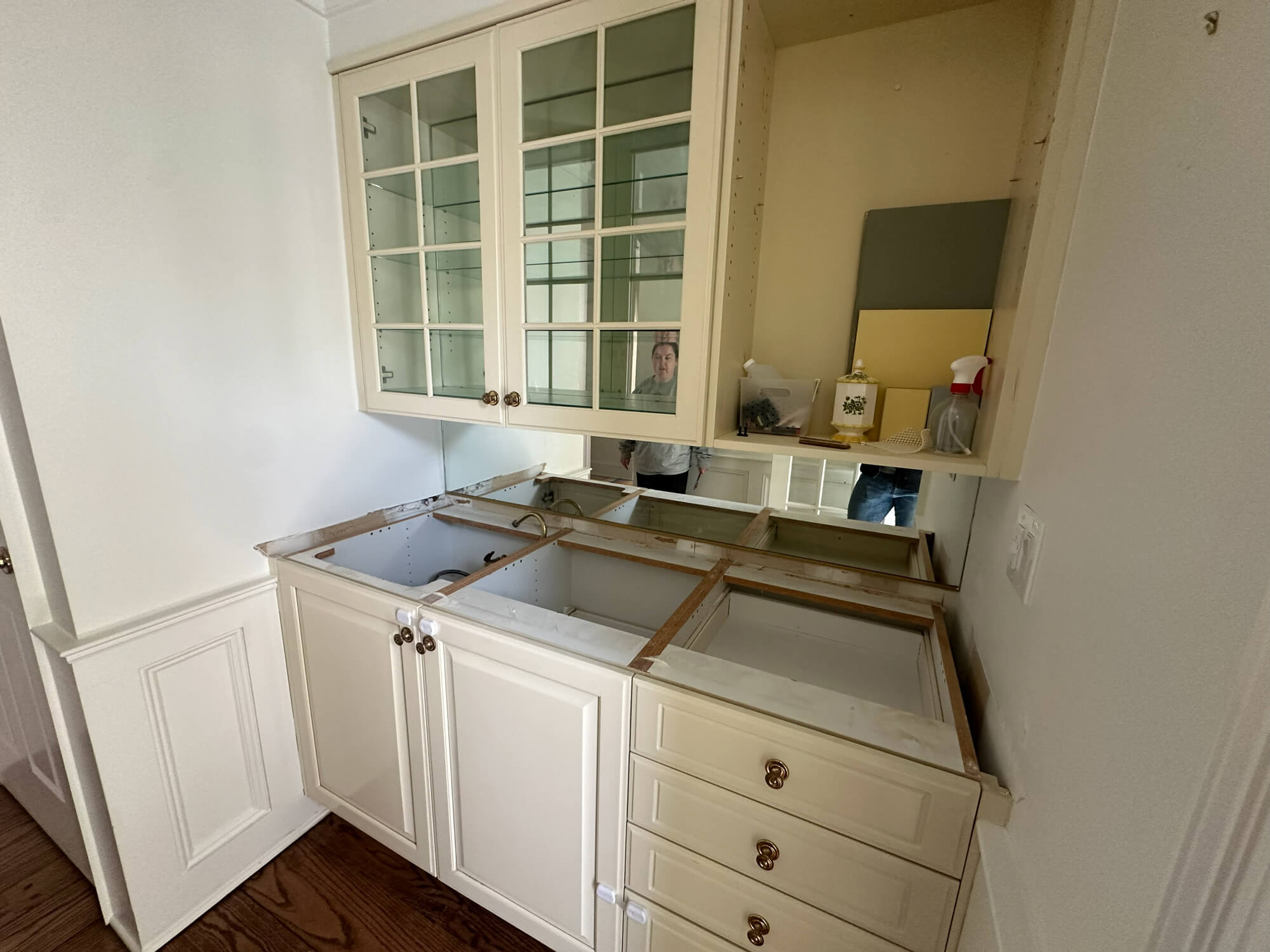 A white kitchen or butler's pantry area under renovation with upper glass-front cabinets, lower cabinets missing the countertop, and a mirrored backsplash.