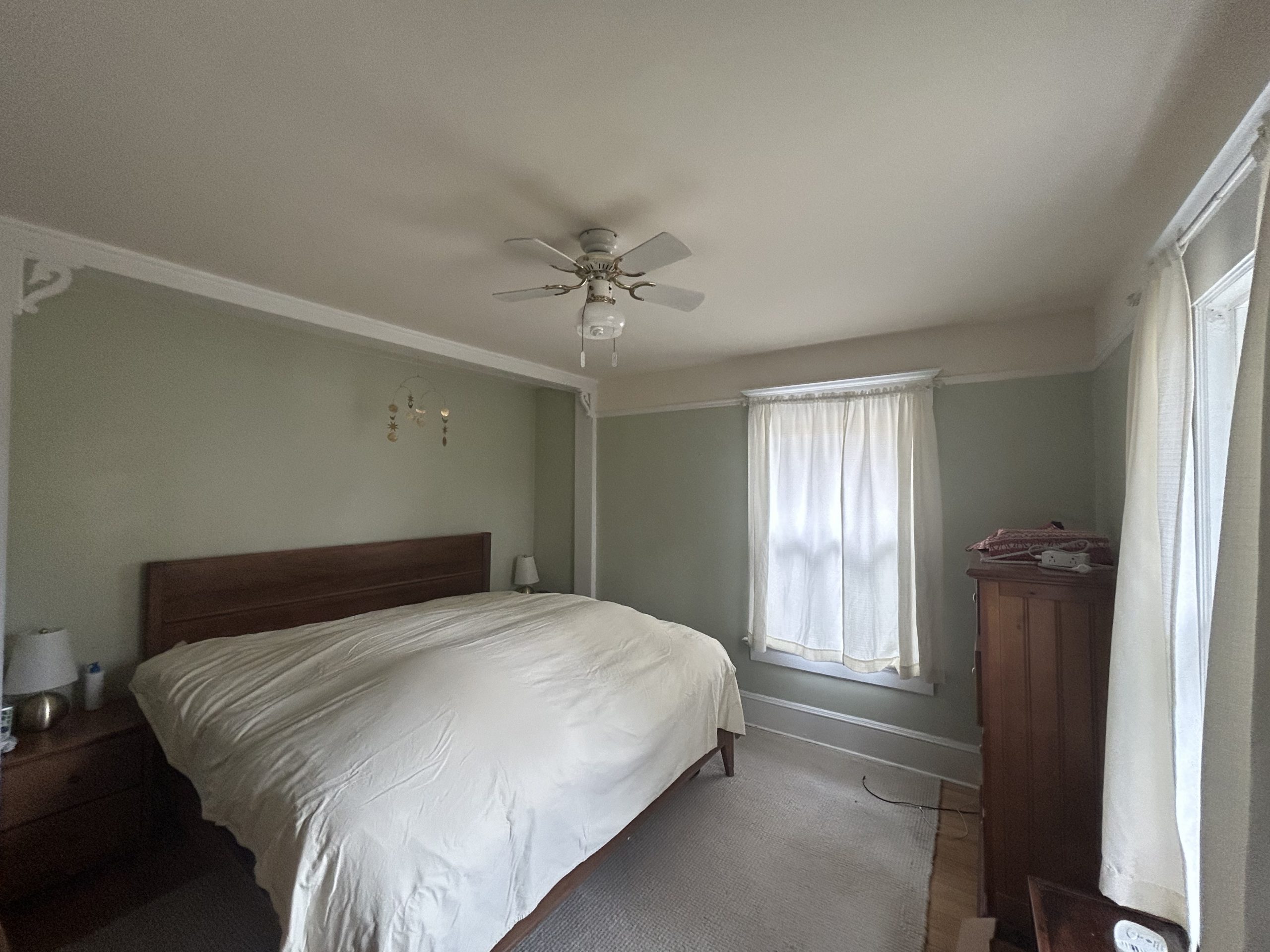 Home Improvement interior painting of a cozy bedroom with light sage green walls, white crown molding, wood furniture, and a ceiling fan.