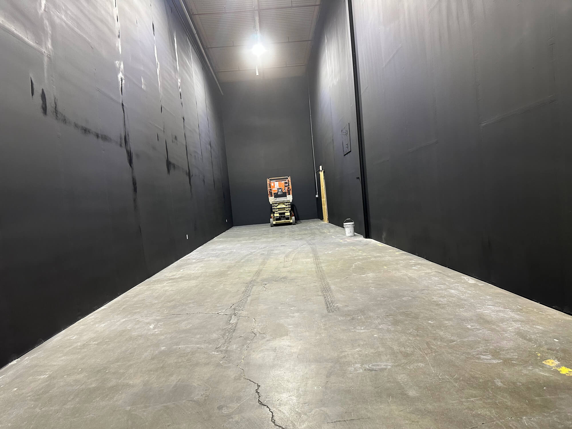 A wide-angle view of a large, empty industrial space or soundstage with concrete flooring and walls newly painted black, with a scissor lift parked in the distance.