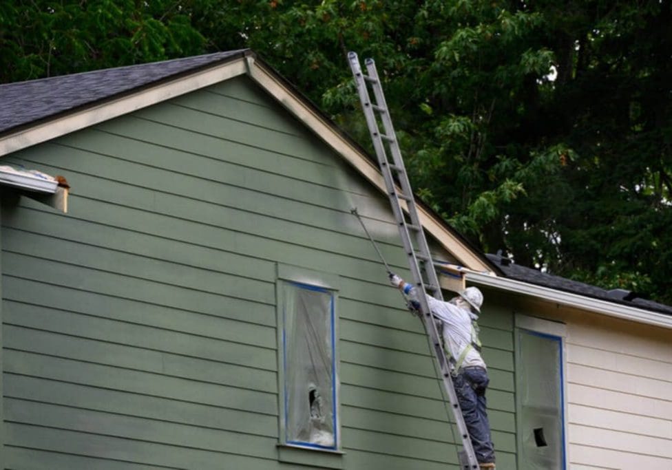 Professional painter on a ladder spraying exterior siding, illustrating how long professional exterior painting typically lasts.