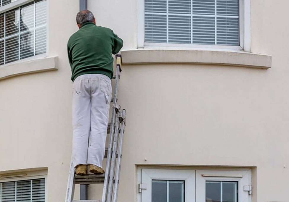 Painter on a ladder working on the exterior of a home, demonstrating how spring weather conditions affect exterior painting projects.