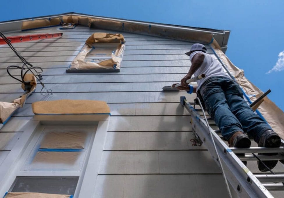 Painter on a ladder rolling exterior siding on a residential home on a sunny day, showing the pros and cons of exterior painting in summer in Warwick, NY.