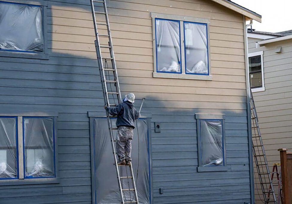 Painter on a ladder spraying exterior siding on a two-story home in Warwick, NY, with windows masked and ready for an exterior painting project.