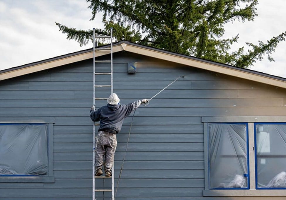 Painter on a ladder spraying exterior siding on a residential home, showing the active work involved in a summer vs. spring exterior painting project.